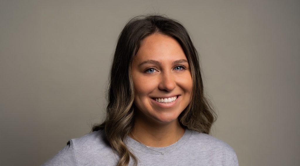 Centered portrait of Cleary designer Emily Perebzak smiling wearing a light gray tshirt standing in front of a darker gray background